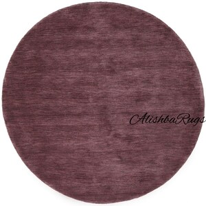 May include: A round rug in a muted burgundy color. The rug has a textured surface and a simple design. The text "Alishba Rugs" is printed on the rug.