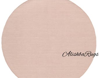 May include: A round, blush-colored rug with a subtle texture. The rug is a solid color and has the text "Alishba Rugs" in a cursive font on the lower right side. The rug is a soft, neutral piece.
