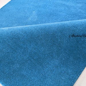 May include: A close-up of a folded, teal-colored rug. The rug has a soft, plush texture and a solid color. The brand name "Atishba Rugs" is visible in the bottom right corner.