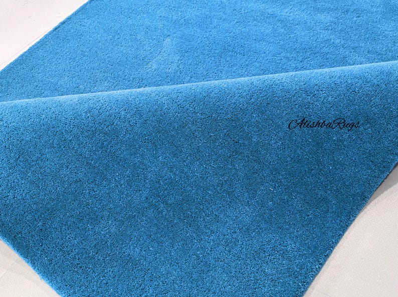 May include: A close-up of a folded, teal-colored rug. The rug has a soft, plush texture and a solid color. The brand name "Atishba Rugs" is visible in the bottom right corner.