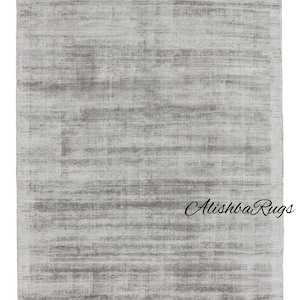 Könnte beinhalten: Ein rechteckiger, hellgrauer Teppich mit strukturiertem Aussehen. Der Teppich hat ein subtiles, horizontales Muster und der Text "Alishba Rugs" in kursiver Schrift ist unten rechts sichtbar.