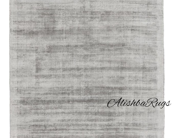 Puede incluir: Una alfombra rectangular gris claro con una apariencia texturizada. La alfombra tiene un patrón horizontal sutil y el texto "Alishba Rugs" en cursiva es visible en la esquina inferior derecha.