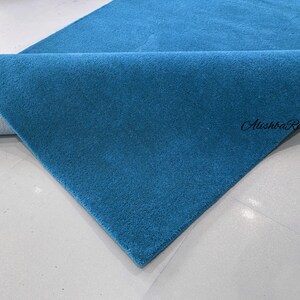 May include: A folded, teal-colored rug with a soft, textured surface. The rug is displayed on a white surface, with the brand name "Alishba Rugs" visible in the corner.
