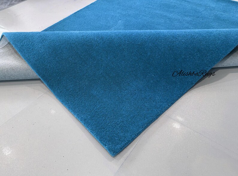 May include: A folded, teal-colored rug with a soft, textured surface. The rug is displayed on a white surface, with the brand name "Alishba Rugs" visible in the corner.