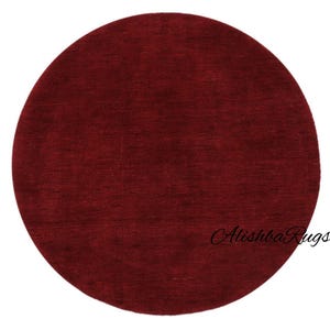 May include: A round, burgundy-colored rug with a textured surface. The rug has a solid color and a simple design. The text "Alishba Rugs" is written in cursive on the bottom right of the rug.