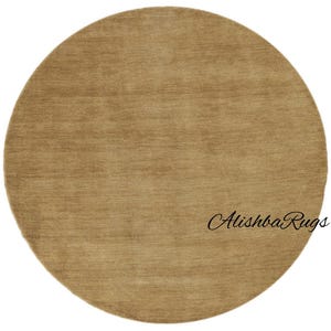 May include: A round, tan-colored rug with a soft, textured surface. The rug has a simple, solid design and is suitable for various interior styles. The text "Alishba Rugs" is written in cursive on the rug.