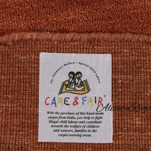May include: Close-up of a textured, rust-colored carpet with a frayed edge. A white label displays the text "CARE & FAIR" and a message about supporting child welfare. The carpet is handmade.
