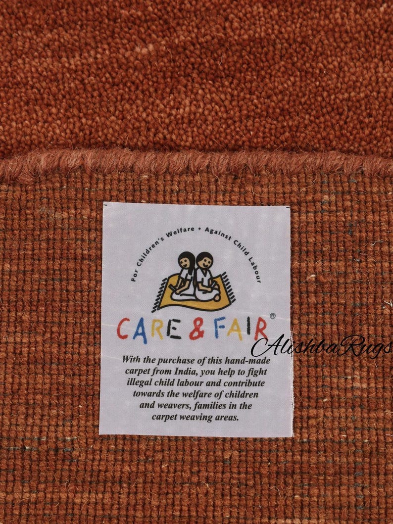 May include: Close-up of a textured, rust-colored carpet with a frayed edge. A white label displays the text "CARE & FAIR" and a message about supporting child welfare. The carpet is handmade.