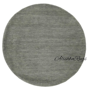 May include: A round, gray rug with a textured surface. The rug is a solid color and appears to be made of a soft material. The text "Alishba Rugs" is printed on the rug.