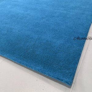 May include: A rectangular, solid blue rug with a soft, textured surface. The rug is a vibrant shade of turquoise and is placed on a light-colored surface. The text "Atishba Rugs" is visible in the image.
