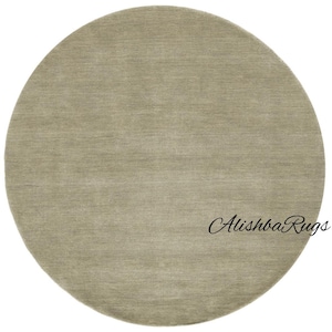 May include: A round, neutral-toned rug with a soft, textured surface. The rug is a solid color, with a subtle variation in tone, and is suitable for various interior design styles. The rug has the text "Alishba Rugs" in the bottom right corner.
