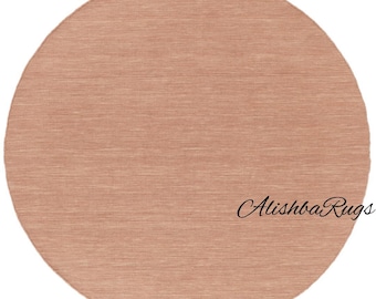 May include: A round, peach-colored rug with a textured surface. The rug has a simple, minimalist design and is suitable for various interior styles. The text "Alishba Rugs" is written in cursive on the rug.