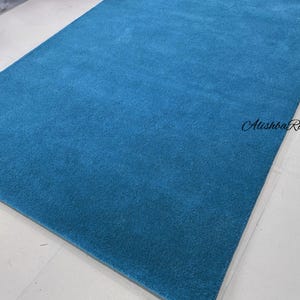 May include: A rectangular, solid teal rug. The rug has a soft, plush texture and is displayed on a white tiled floor. The text "Atishba Rugs" is visible in the upper right corner.