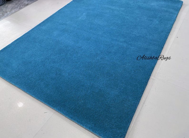May include: A rectangular, solid teal rug. The rug has a soft, plush texture and is displayed on a white tiled floor. The text "Atishba Rugs" is visible in the upper right corner.