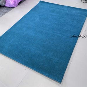 May include: A rectangular, teal-colored rug. The rug has a soft, plush texture and a solid color design. The rug is displayed on a white tiled floor. The text "Alishba Rugs" is in the bottom right corner.