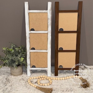 May include: Two wooden ladder picture frames, one whitewashed and one brown, each with three rectangular openings for photos. The frames are standing on a white fluffy surface with a string of wooden beads and a tassel.