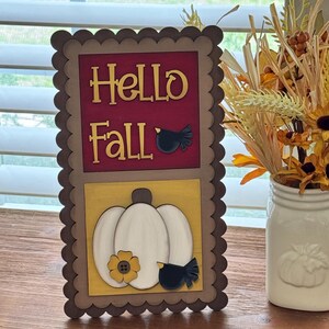 May include: A wooden fall-themed sign with the words "Hello Fall" in gold lettering on a burgundy background. Below, a white pumpkin with a yellow flower and a black bird is displayed. The sign has a scalloped edge and is on a wooden surface.