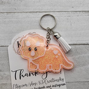 May include: A pink glitter keychain with a white outline of a cartoon dinosaur. The keychain has a white tassel and a silver key ring. The tag attached to the keychain says "Thank you!" and includes the Etsy shop name and social media handles.