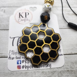 May include: A black and gold honeycomb charm with a black and gold bead, a white bead with the text "Be Kind" and a gold glitter bead. The charm is attached to a black cord.