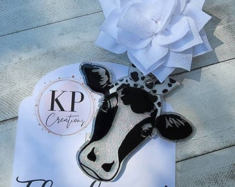 Puede incluir: Un llavero de cabeza de vaca en blanco y negro con detalles de purpurina. El llavero está unido a una flor de tela blanca con un clip negro. El llavero está sobre una tarjeta blanca con el texto "KP Creations" y "¡Gracias!"  Etsy.com/shop/KPCreationsky. También encuéntranos en Facebook.