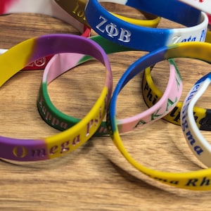 May include: A collection of colorful silicone wristbands with Greek letters and names, including "Alpha Psi", "Zeta Phi Beta", "Omega", "Kappa", and "Phi Sigma Rho".