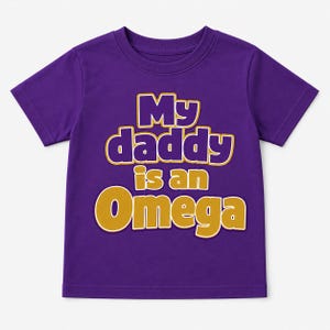 May include: A purple short-sleeved t-shirt with the text "My daddy is an Omega" in gold and white lettering. The shirt is a solid purple color and is likely for a child.