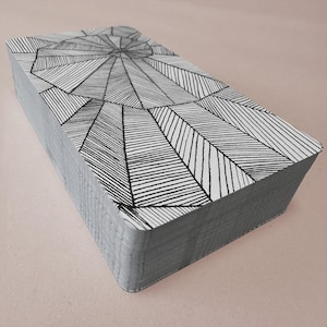 K&ouml;nnte beinhalten: Ein Kartenspiel mit einem geometrischen Schwarz-Wei&szlig;-Design. Die Karten haben graue Kanten und ein Muster aus Linien, die von einem zentralen Punkt ausgehen. Die Karten sind ordentlich gestapelt und zeigen das detaillierte Design auf der obersten Karte.
