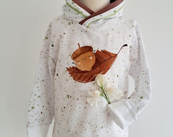 Könnte beinhalten: Weißer Kapuzenpullover mit braunem und grünem Besatz. Der Pullover hat eine braune und grüne Eichel mit einem lächelnden Gesicht auf einem braunen Blatt. Der Hintergrund ist weiß mit braunen und grünen Spritzflecken.