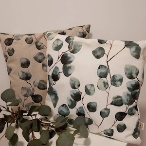Op de afbeelding: Twee vierkante kussens met een botanisch eucalyptusblad-ontwerp. Eén kussen heeft een beige achtergrond, de andere een witte achtergrond. Beide hebben groene en grijze bladeren met bruine stelen, wat een natuurlijke, rustgevende uitstraling creëert.