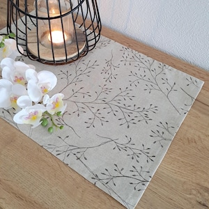 Op de afbeelding: Een beige tafelkleed met een delicaat zwart bloemenpatroon. De tafelkleed is gemaakt van een linnenachtige stof en heeft een subtiel, herhalend patroon van kleine takken en bladeren.