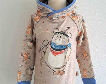Könnte beinhalten: Ein beiger Kapuzenpullover mit blauem Besatz. Der Pullover zeigt einen Comic-Bären, der ein blaues Bandana und ein rotes Stirnband trägt. Der Bär hält einen Speer und Federn. Der Hintergrund ist beigefarben mit einem Muster aus Federn und Pfeilen.