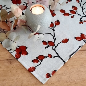 Könnte beinhalten: Ein weißer Tischläufer mit einem roten Beeren- und Astdesign. Ein grauer Kerzenhalter mit einer brennenden Kerze steht auf dem Läufer.