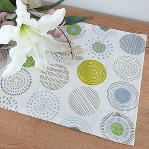 Op de afbeelding: Een witte tafelrunner met een geometrisch patroon van groene, grijze en blauwe cirkels. De cirkels hebben verschillende texturen en ontwerpen, waaronder swirls, strepen en stippen.