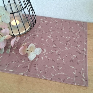 Könnte beinhalten: Ein rosa Tischläufer mit einem weißen Blumenmuster. Der Läufer ist aus einem weichen, gewebten Stoff gefertigt und hat einen dezenten Glanz.