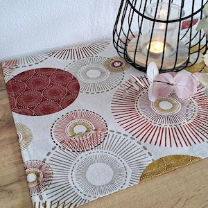 Könnte beinhalten: Dekorativer Tischläufer mit einem sich wiederholenden Muster aus roten, goldenen und grauen konzentrischen Kreisen auf weißem Grund. Ein schwarzer Draht-Kerzenhalter mit einer brennenden Kerze und Blumen befinden sich auf dem Tischläufer.