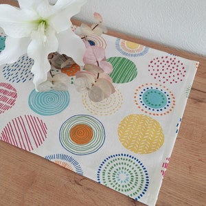 Könnte beinhalten: Ein weißer Tischläufer mit einem farbenfrohen Muster aus Kreisen. Die Kreise sind mit verschiedenen Mustern gefüllt, darunter Streifen, Punkte und Spiralen. Der Läufer ist perfekt, um jeder Tischdekoration einen Hauch von Verspieltheit zu verleihen.