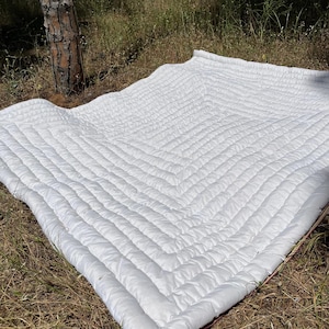 Puede incluir: Una manta blanca acolchada con un patrón geométrico, extendida sobre el suelo. La manta tiene una textura suave y esponjosa y una estética limpia y minimalista. Perfecta para uso al aire libre.