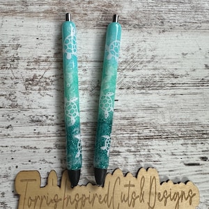 May include: Two turquoise and teal glitter pens with white sea turtle designs. The pens have black tips and silver accents. The pens are resting on a wooden sign that reads "Torris Inspired Cuts & Designs".