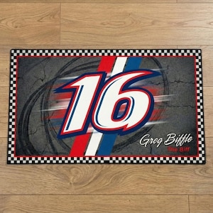 May include: Rectangular doormat with a grey background featuring tyre tracks and the number 16 in white, red, and blue. The name "Greg Biffle" is printed in cursive. The mat has a black and white chequered border.