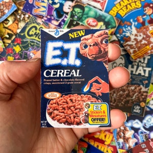 May include: A miniature cereal box for E.T. Cereal, featuring the E.T. character on a navy blue background. The box advertises peanut butter and chocolate-flavored cereal. The box is held in a hand, with other cereal boxes in the background.