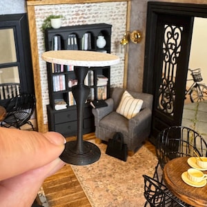 May include: A miniature black table with a speckled beige tabletop is held in a hand. The table has a decorative, slender pedestal base and a round, flat base. The table is part of a dollhouse interior scene with a bookshelf, armchair, and other furniture.