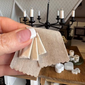 May include: A beige linen fabric with a scalloped edge and a folded section of the same fabric on top. The fabric is being held by a hand.