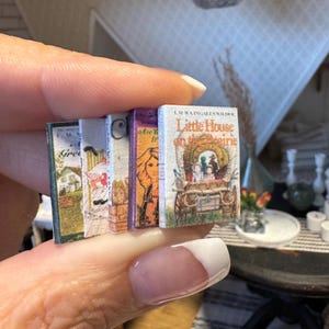 May include: A collection of miniature books, held in a hand. The books have colorful covers with titles like "Little House on the Prairie" and "Are You My Mother?" The books are small, likely for a dollhouse or miniature display.