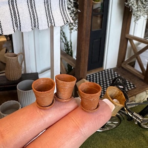 May include: Three small terracotta pots with matching saucers. The miniature pots are held between fingers, with a miniature bicycle and a dollhouse porch in the background. The pots are empty and ready for planting.
