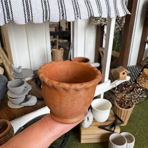 Op de afbeelding: Een kleine bloempot van terracotta met een geschulpte rand. De pot wordt in een hand gehouden, met op de achtergrond miniatuurdecoraties zoals een vogelbeeldje, kopjes en een vogelhuisje, wat een miniatuurtuin thema suggereert.