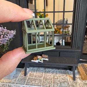 May include: A miniature green greenhouse with a wooden frame and glass panels. The greenhouse is being held in a person's hand.