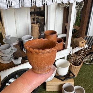 Puede incluir: Una pequeña maceta de terracota con un borde festoneado. La maceta sin esmaltar está sostenida por un dedo, destacando su tamaño. Otros artículos de cerámica en miniatura son visibles en el fondo, incluyendo tazas, platillos y macetas adicionales.