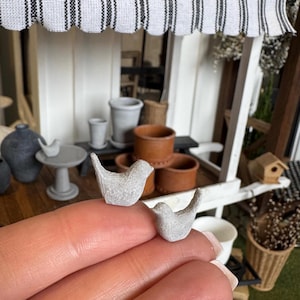 Peut inclure: Deux figurines d'oiseaux gris miniatures. Les oiseaux sont posés sur une surface, avec une scène de jardin miniature en arrière-plan. La scène comprend des pots miniatures, une table et un petit nichoir, le tout dans une palette de couleurs neutres.