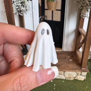 May include: A small, white, three-dimensional ghost figurine with a smooth, matte finish. The ghost features a rounded head with two hollow, oval-shaped eye openings and a draped, flowing body that flares out at the bottom, creating a classic spectral shape.