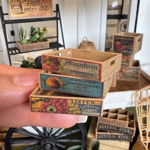 May include: A stack of three miniature wooden crates featuring vintage-style labels. The top crate displays Mountain Cherries, the middle crate shows Colorado Peaches, and the bottom crate features Listen Brand California Grapes with colourful fruit illustrations.